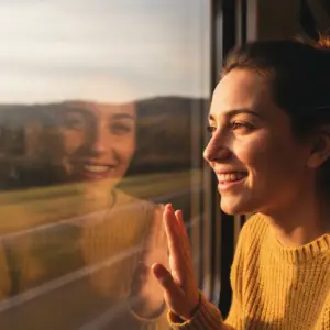 Close-up photograph of a person smiling genuinely while looking out of a train window during golden hour, blurred scenery passing by, soft light illuminating their face, conveying a sense of hope and forward movement.